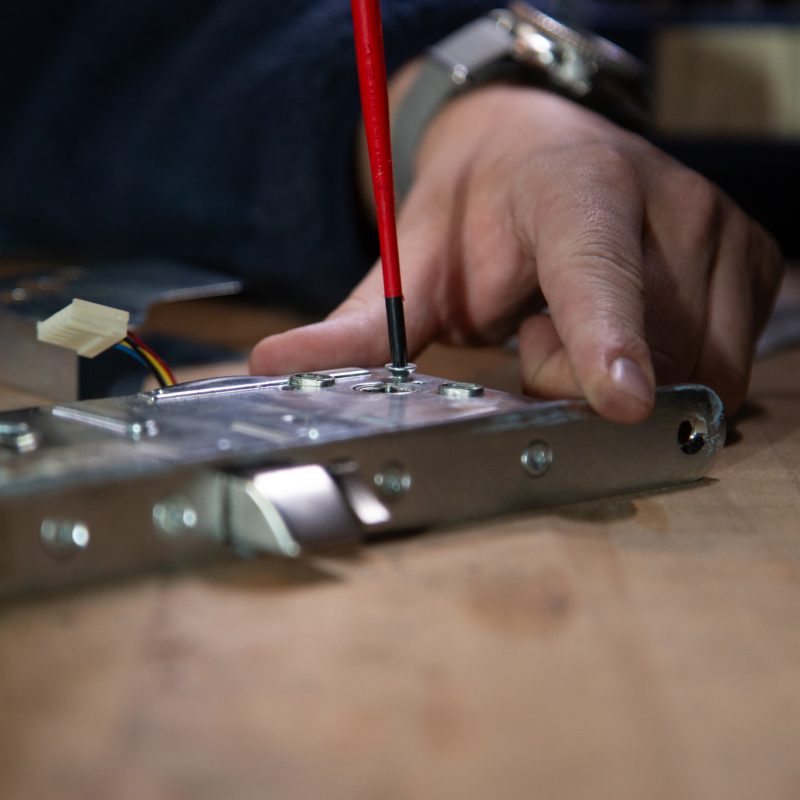 En persons hånd bruger en skruetrækker med rødt håndtag til at spænde en skrue på en metalkomponent med ledninger, der hviler på træ - hvilket tyder på montage- eller reparationsarbejde, typisk i et låsesmed danmark københavn låseservice akut-scenarie.